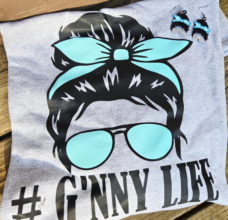 May include: A grey t-shirt featuring a graphic of a woman's face with sunglasses and a headband. The sunglasses and headband are light blue. The text "# G'nny Life" is printed on the shirt. Matching earrings are also visible.