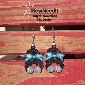 May include: A pair of handmade earrings depicting a black hair bun with a light blue bow and silver sunglasses. The earrings are displayed on a red and brown wooden surface. The text "I SewNeedIt" is visible.