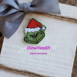 Könnte beinhalten: Eine gestickte Grinch-Figur in Grün und Weiß, die einen roten Weihnachtsmann-Hut trägt. Die Stickerei befindet sich auf einem weißen Holzhintergrund mit einer grauen Schleife und Schnur. Der Text "ISewNeedIt" und "Digitaler Download" befindet sich unter der Stickerei.