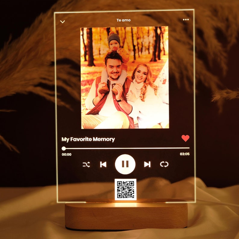May include: A clear acrylic photo frame with a family photo inside. The frame has a music player interface with buttons for play, pause, rewind, and fast forward. The text "My Favorite Memory" is displayed above the music player. The frame is lit from below with a warm white light.