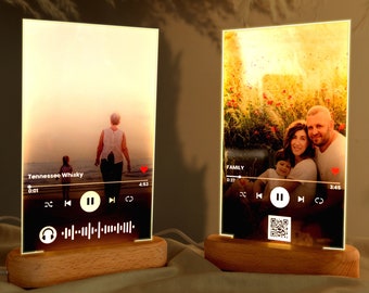 Pode incluir: Dois quadros de fotos de acrílico iluminados com bases de madeira. Cada quadro exibe uma foto e uma interface de reprodutor de música com títulos de músicas, "Tennessee Whiskey" e "FAMILY", e controles de reprodução. O quadro à esquerda mostra uma foto de uma pessoa caminhando em uma praia ao pôr do sol. O quadro à direita mostra uma foto de uma família de três pessoas em um campo de flores.