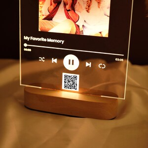 May include: A clear acrylic photo frame with a wooden base. The frame has a digital music player interface with a photo, the text "My Favorite Memory", and a QR code. The interface shows a play button, a pause button, and a rewind button.