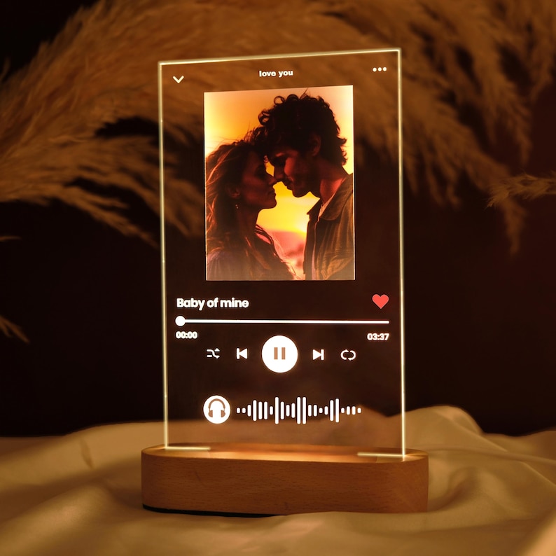 May include: A clear glass lamp with a wooden base. The lamp features a photo of a couple, a music player interface with the text "Baby of mine", and a heart icon. The music player interface shows a play button, a pause button, a rewind button, and a fast forward button. The lamp also has a headphone icon and a waveform.