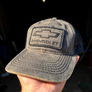 May include: Gray Chevrolet trucker hat with a black mesh back. The front panel features the Chevrolet logo and the brand name in black. The hat has a curved brim and a distressed look.