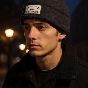 May include: A dark gray beanie with a white rectangular patch featuring the Chevrolet logo. The hat is worn by a person wearing a black jacket. The background is blurred with streetlights.