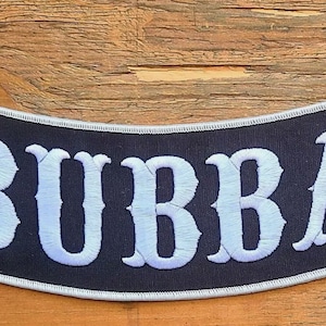 May include: Black, arched patch with the name "BUBBA" embroidered in white, stylized lettering. The patch has a white border and is set against a wooden background.