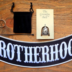 May include: A black and white embroidered patch with the word "Brotherhood" in white lettering. The patch is on a wooden surface with a silver motorcycle bell, a black velvet pouch, and a silver ring.