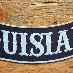May include: A black and white embroidered patch with the word "LOUISIANA" in white lettering on a black background. The patch is curved and has a white border.