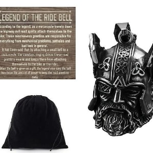 May include: A silver-colored motorcycle ride bell pendant in the shape of a Viking helmet. The pendant has intricate details and a loop for attaching to a chain. A black drawstring bag and a wooden sign with text are also in the image.