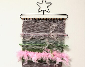 May include: A woven wall hanging with pink and green yarn, featuring pink feathers and wooden dowels. The hanging is attached to a black metal star-shaped hook.