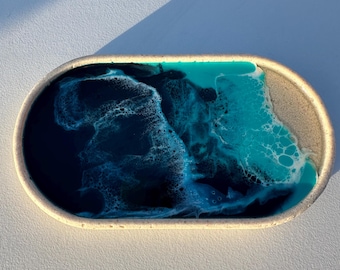 May include: An oval-shaped tray with a white rim and a blue and black resin design that resembles an ocean wave.