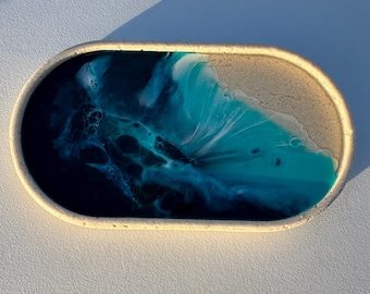 May include: An oval-shaped tray with a white concrete exterior and a resin interior depicting an ocean scene. The resin is blue, teal, white, and black, resembling waves crashing on a sandy beach.
