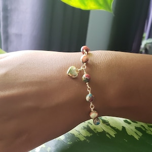 May include: A gold bracelet with a heart charm and colorful beads. The bracelet is on a person's wrist.