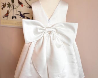 May include: A white satin dress with a large bow on the back, displayed on a mannequin. The dress has a V-neck and a full skirt. The bow is the focal point, adding a touch of elegance. The dress is suitable for special occasions.