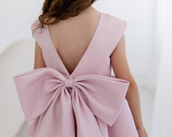 May include: A light pink sleeveless dress with a large bow in the back. The dress has a deep V-neckline and a fitted bodice.
