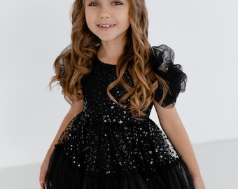 Könnte beinhalten: Ein junges Mädchen trägt ein schwarzes Tüllkleid mit Pailletten. Das Kleid hat einen gerüschten Rock und Puffärmel.