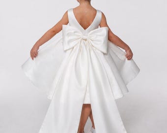 May include: A sleeveless ivory satin dress featuring a deep V-neckline on the back and a large, structured bow at the waist. The dress has a flared skirt with two long, wide fabric panels extending from the bow down to the floor.