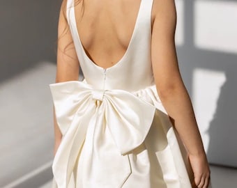 May include: Ivory-colored dress with a large bow at the back. The dress features a scoop back and a full skirt. The bow is a prominent detail, adding a touch of elegance. The dress appears to be made of a smooth, satin-like fabric.