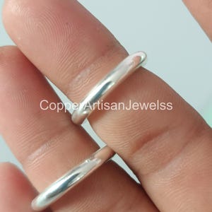 May include: A close-up of two silver rings. The rings are smooth and shiny, with a rounded band. The text "Copper Artisan Jewelss" is visible in the image. The rings are likely made of sterling silver and are a minimalist design.