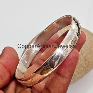 May include: A silver bangle bracelet with a smooth, polished finish. The bracelet is open and has a slightly rounded shape.