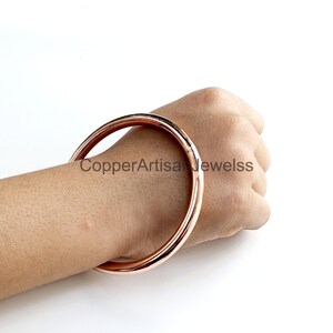 Puede incluir: Un brazalete de oro rosa. El brazalete liso y circular se muestra en la muñeca de una persona. El texto "CopperArtisanJewelss" es visible en la imagen.