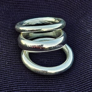 May include: Three silver rings stacked on top of each other. The rings are thick and have a smooth, polished finish. The rings are displayed on a dark blue textured surface. The rings are likely jewelry.
