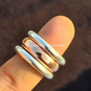 May include: Close-up of three stacked rings. The top and bottom rings are silver, while the middle ring is copper. The rings are cylindrical and smooth, with a slight curve. The rings are on a finger, with a brown background.