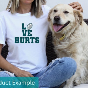 K&ouml;nnte beinhalten: Wei&szlig;es T-Shirt mit einem gr&uuml;nen Football und dem Text "LOVE HURTS" auf der Vorderseite. Eine Person tr&auml;gt das T-Shirt und sitzt mit einem Hund.
