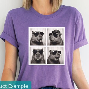 May include: A purple t-shirt featuring a square graphic of four capybaras in a photo booth style. Each capybara wears sunglasses, and one has a hat. The shirt is a solid color and has rolled-up sleeves.