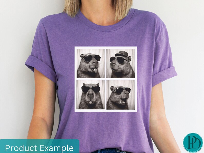 May include: A purple t-shirt featuring a square graphic of four capybaras in a photo booth style. Each capybara wears sunglasses, and one has a hat. The shirt is a solid color and has rolled-up sleeves.