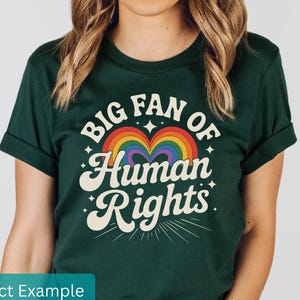 May include: Dark green t-shirt with the text "BIG FAN OF Human Rights" in a retro font, featuring a rainbow heart design. The shirt has rolled-up sleeves and a crew neck.