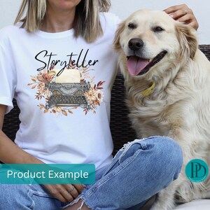 May include: White t-shirt with the word "Storyteller" printed in black script. A vintage typewriter with floral accents is printed below the text. The t-shirt is being worn by a person with a dog sitting next to them.
