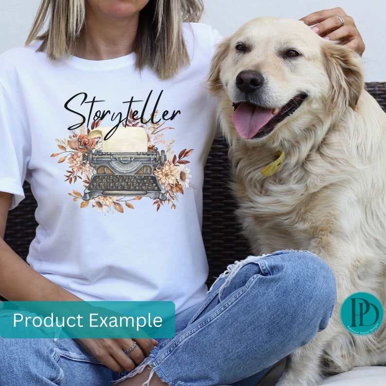 May include: White t-shirt with the word "Storyteller" printed in black script. A vintage typewriter with floral accents is printed below the text. The t-shirt is being worn by a person with a dog sitting next to them.