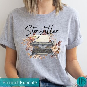 May include: Gray t-shirt with the text "Storyteller" printed in black. The design features a vintage typewriter with a blank sheet of paper and floral accents in shades of brown, orange, and white.