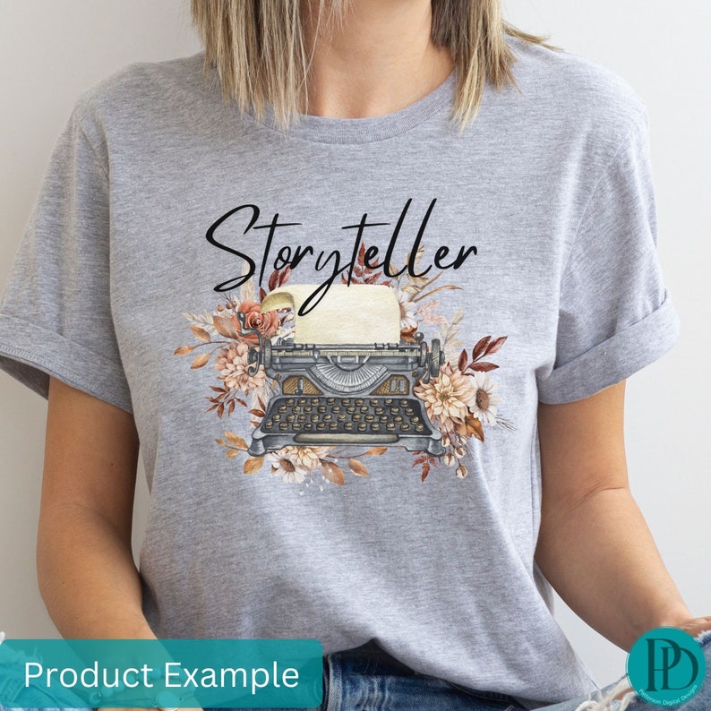 May include: Gray t-shirt with the text "Storyteller" printed in black. The design features a vintage typewriter with a blank sheet of paper and floral accents in shades of brown, orange, and white.