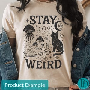 May include: A cream-coloured t-shirt with a black graphic design. The design includes a cat, mushrooms, a jellyfish, and the words "STAY WEIRD." The shirt is worn with a denim jacket. The design also includes stars, a moon, and other mystical elements.
