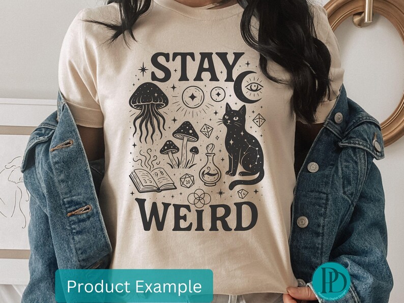 May include: A cream-coloured t-shirt with a black graphic design. The design includes a cat, mushrooms, a jellyfish, and the words "STAY WEIRD." The shirt is worn with a denim jacket. The design also includes stars, a moon, and other mystical elements.