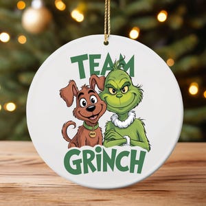 K&ouml;nnte beinhalten: Wei&szlig;er Keramik-Weihnachtsschmuck mit dem Grinch und dem Hund Max. Der Grinch ist gr&uuml;n mit einem wei&szlig;en Fellkragen, und Max ist braun. Die Worte "Team Grinch" sind gr&uuml;n gedruckt.
