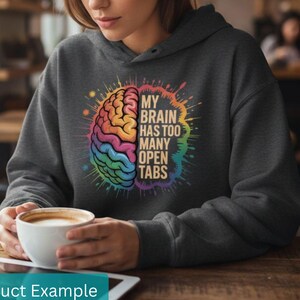 May include: Dark gray hoodie with a colorful brain graphic. The graphic features a rainbow-colored brain with the text "MY BRAIN HAS TOO MANY OPEN TABS". The hoodie is worn by a person holding a white coffee cup.