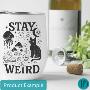 May include: White stemless wine tumbler with a black graphic design featuring a cat, mushrooms, and the words "STAY WEIRD". A bottle of wine, a corkscrew, and a cork are in the background.