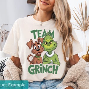 K&ouml;nnte beinhalten: Cremefarbenes T-Shirt mit einem gr&uuml;nen "TEAM GRINCH"-Aufdruck, der den Grinch und einen Hund zeigt. Der Hund hat ein goldfarbenes Halsband. Das Shirt ist l&auml;ssig.