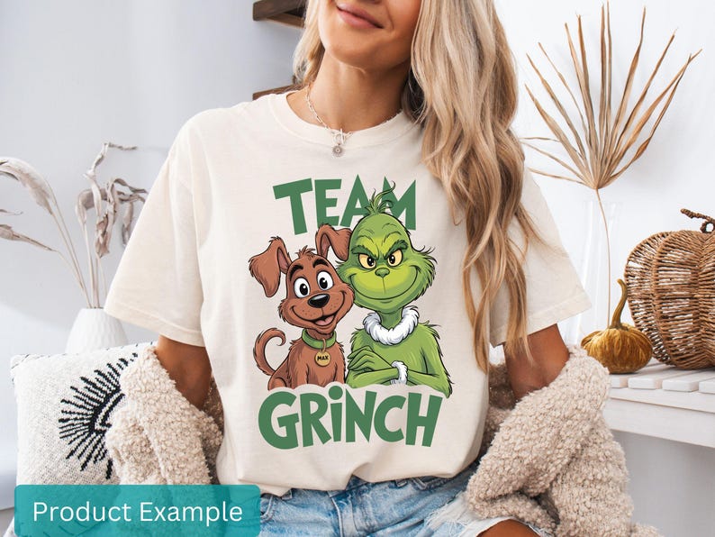 K&ouml;nnte beinhalten: Cremefarbenes T-Shirt mit einem gr&uuml;nen "TEAM GRINCH"-Aufdruck, der den Grinch und einen Hund zeigt. Der Hund hat ein goldfarbenes Halsband. Das Shirt ist l&auml;ssig.