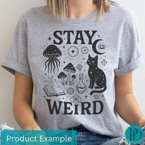May include: A grey t-shirt featuring a black graphic with the words "STAY WEIRD". The design includes a black cat, mushrooms, a jellyfish, a potion bottle, and other mystical symbols.