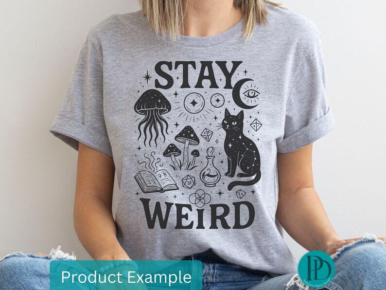 May include: A grey t-shirt featuring a black graphic with the words "STAY WEIRD". The design includes a black cat, mushrooms, a jellyfish, a potion bottle, and other mystical symbols.