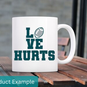 K&ouml;nnte beinhalten: Eine wei&szlig;e Keramik-Kaffeetasse mit einem blaugr&uuml;nen Design, auf dem "Love Hurts" mit einer Fu&szlig;ballgrafik steht.