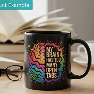 May include: Black ceramic mug with a colorful brain graphic and the text "MY BRAIN HAS TOO MANY OPEN TABS". The mug is on a wooden surface with a book, glasses, and a cup of coffee in the background.