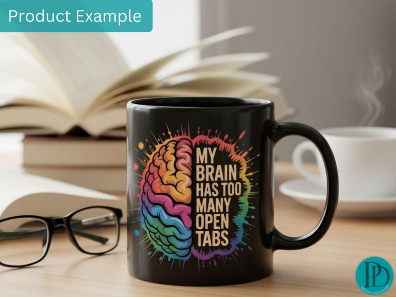 May include: Black ceramic mug with a colorful brain graphic and the text "MY BRAIN HAS TOO MANY OPEN TABS". The mug is on a wooden surface with a book, glasses, and a cup of coffee in the background.