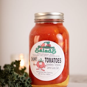 Peut inclure: Un bocal en verre rempli de tomates rouges et d'un liquide jaune. Le bocal porte une étiquette blanche avec le texte "Down Home Salads" et "CHUNKY TOMATOES". L'étiquette indique également les ingrédients : Tomates, Jus de citron, Sel. Le bocal est posé sur une surface blanche.