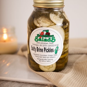 May include: A glass jar filled with sliced dill pickles in a clear brine. The jar has a white label with green text that reads "Down Home Salads", "Salty Brine Pickles", and the ingredients and net weight.