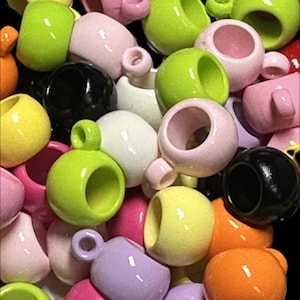 May include: A collection of colorful ceramic beads in various shades, including pink, green, yellow, orange, and black. Each bead has a circular opening and a small handle, resembling miniature cups. The beads are glossy and arranged against a black background.